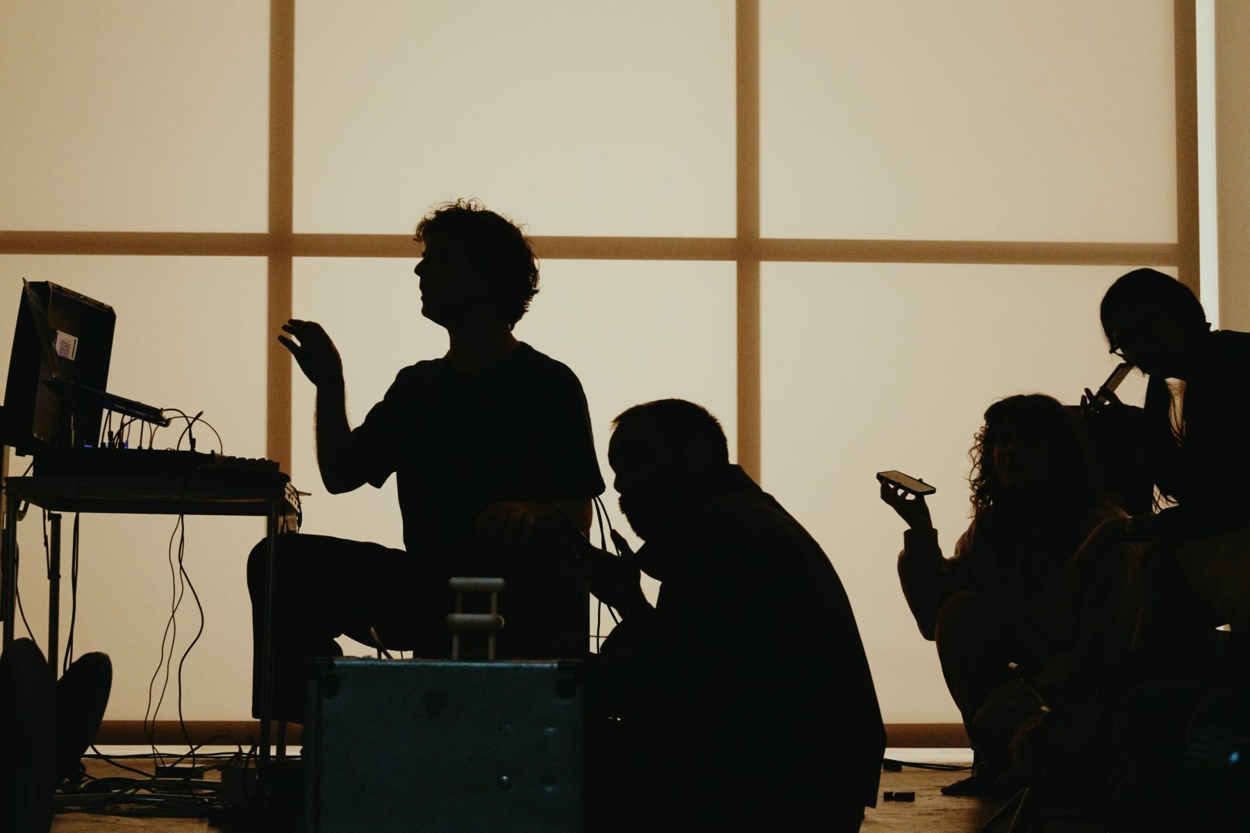 A group of people in silhouetter, one in front of a laptop and modular synth, the others holding mobile devices out in front of them